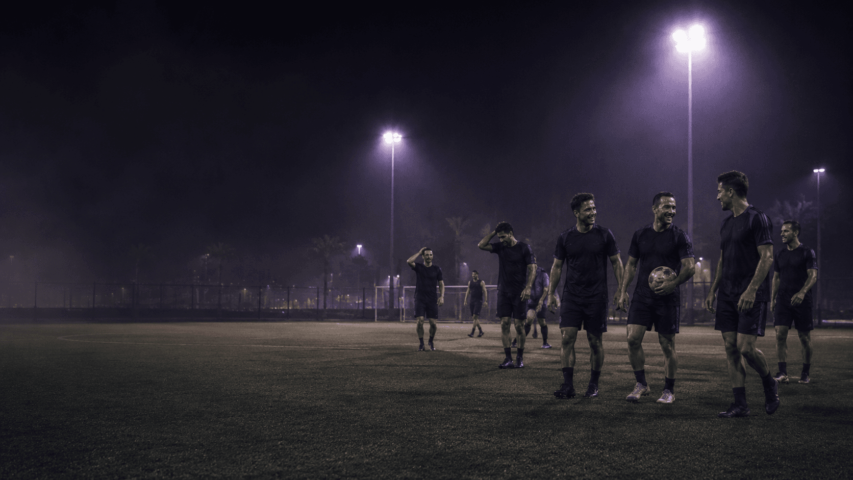 Adult football players walking together after a session on a floodlit pitch